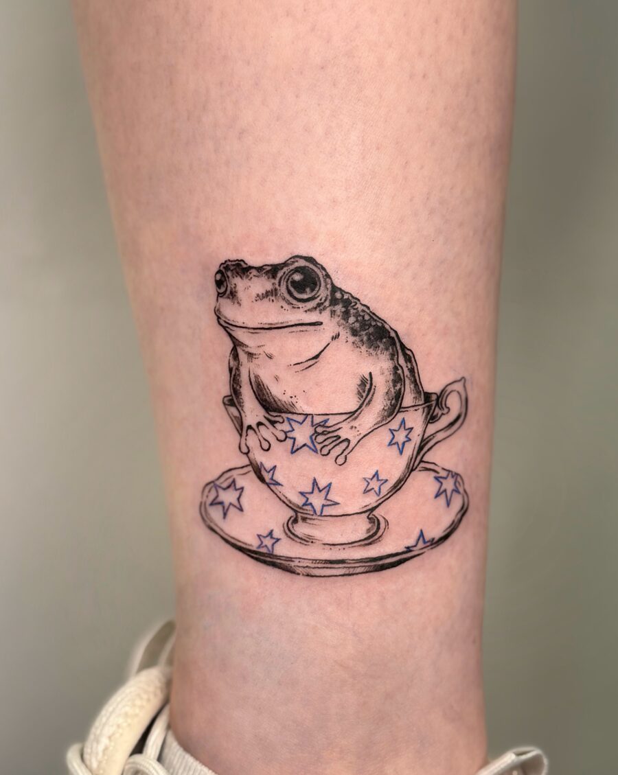 A playful frog nestled in a teacup Tattoo on skin
